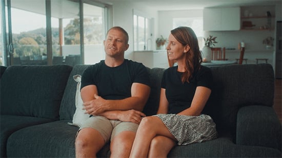 A medium shot of a man and a woman sitting on a dark grey couch in a modern, open-plan living room, looking toward the side of the frame while engaged in conversation.