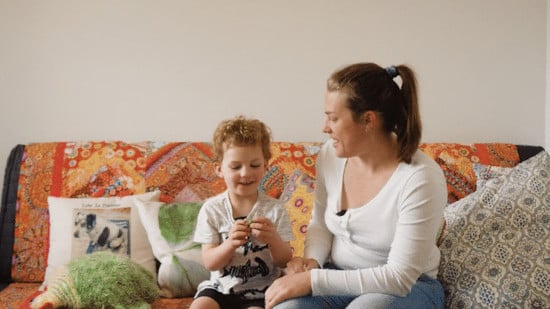 A woman and a young boy sitting together on a vibrant, patterned couch, sharing a smile as the boy plays with a small object in his hands.