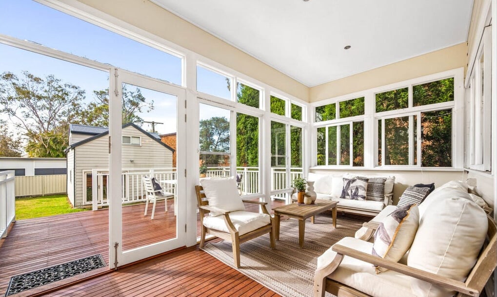 A bright, sun-drenched sunroom featuring white-framed windows and doors that open onto a wooden deck, furnished with comfortable white seating and a neutral area rug.