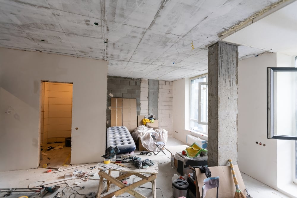 An eye-level shot of a large room in the middle of a renovation, showing exposed concrete ceilings, unfinished block walls, and a variety of construction tools and equipment scattered across the space.