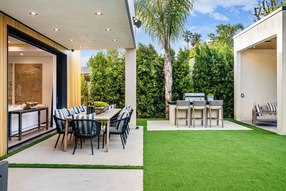 A wide shot of a luxurious, modern backyard featuring a covered patio with an outdoor dining set, an artificial turf lawn, and a built-in outdoor kitchen