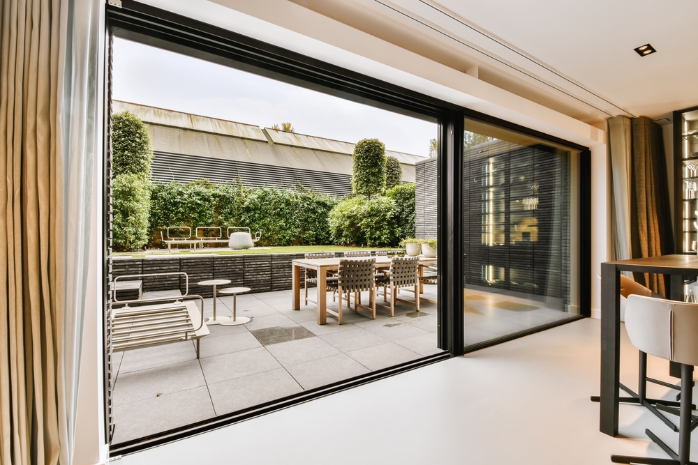 A wide shot from inside a modern home looking through large, black-framed sliding glass doors onto a sleek tiled patio featuring an outdoor dining set and minimalist lounge furniture.