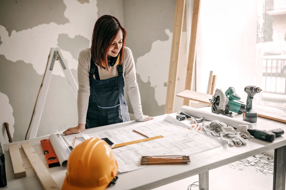A high-angle shot of a female builder in denim overalls smiling as she reviews architectural blueprints on a workbench surrounded by power tools and a yellow hard hat.