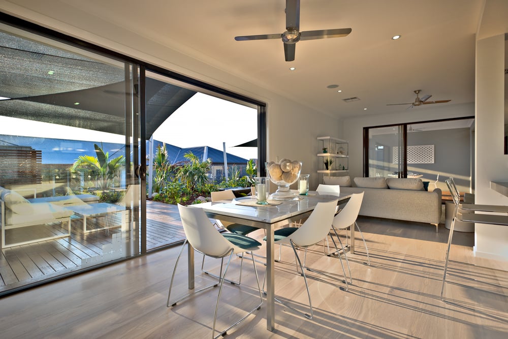 A wide, eye-level shot of a bright, modern interior featuring a dining area and living space with sliding glass doors that open onto a sunlit wooden deck with outdoor seating.