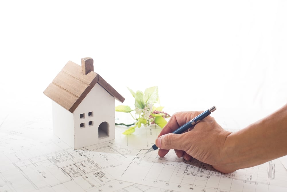 A high-angle, close-up shot of a hand using a mechanical pencil to draft notes on architectural blueprints next to a small, decorative model house.