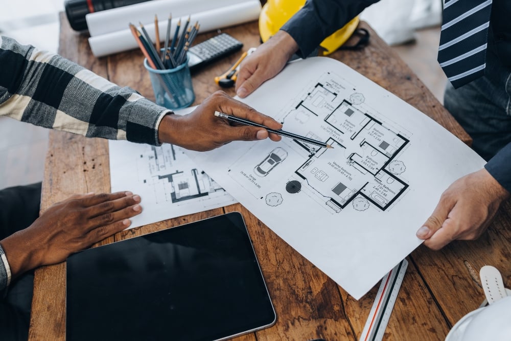 A high-angle, close-up shot of two people reviewing a set of architectural floor plans on a wooden desk, with one person pointing a pencil at a specific room layout.