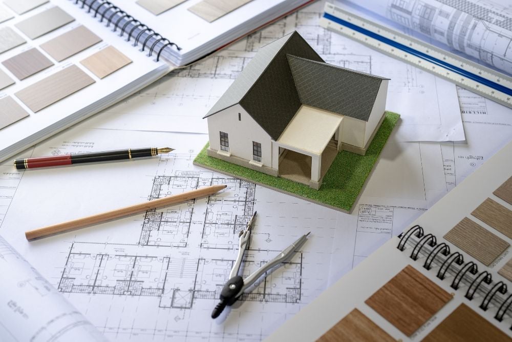 An architectural model of a house sits on a desk surrounded by floor plans, a compass, pens, and material sample books.