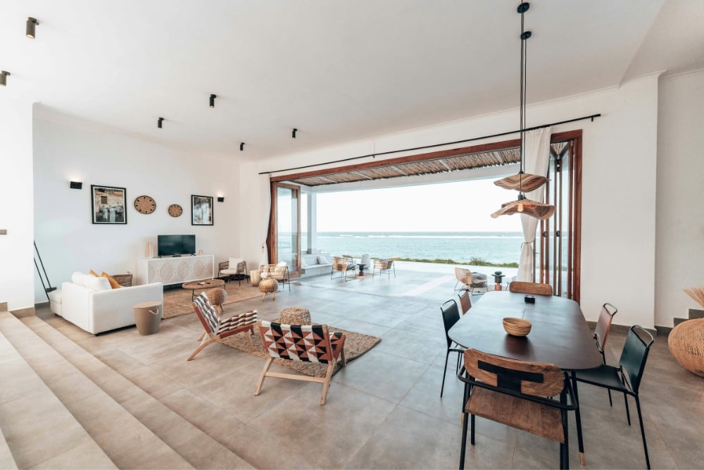 A wide, high-angle shot of a spacious, coastal-style living and dining area with bi-fold doors that open completely to a terrace overlooking the ocean.