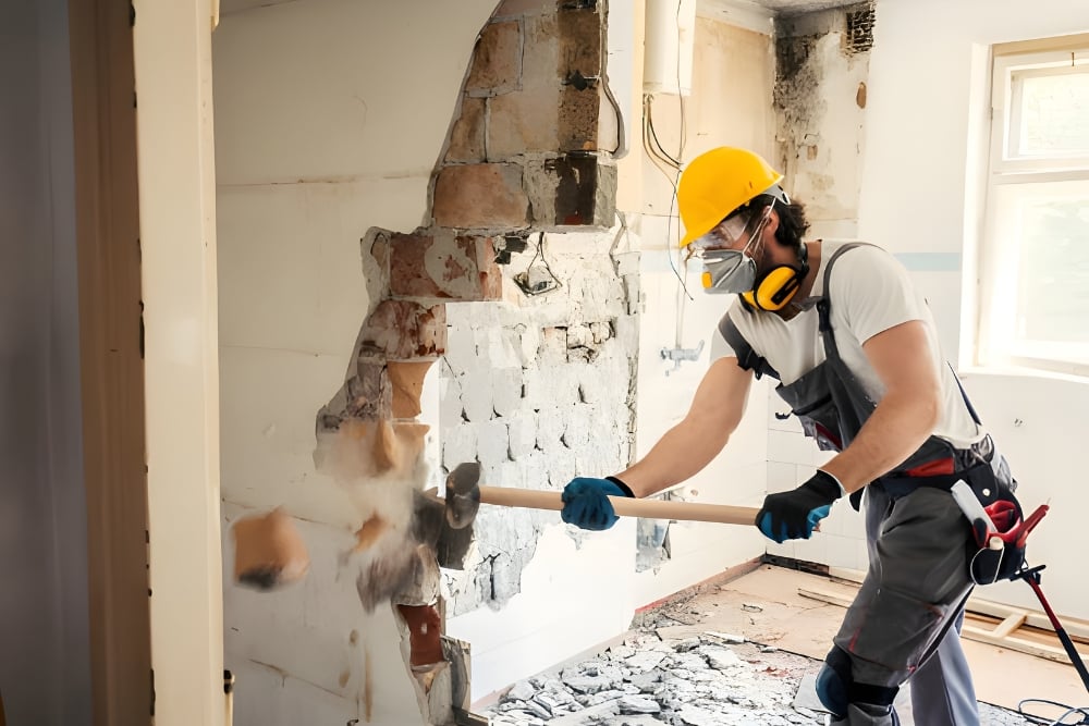 A construction worker wearing a yellow hard hat, respirator, and safety goggles uses a sledgehammer to demolish an interior brick and plaster wall.
