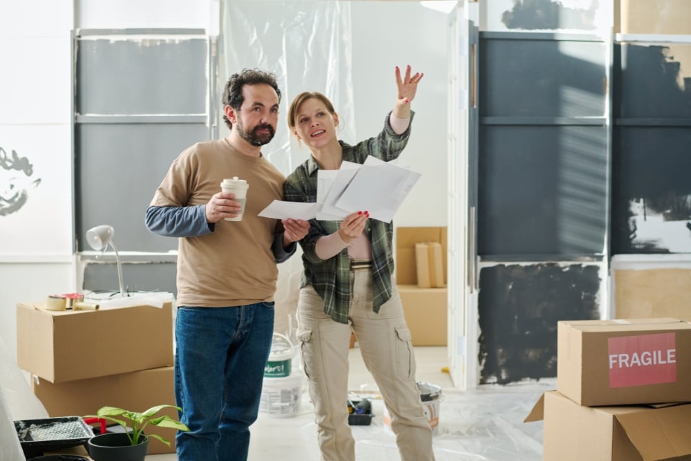 An eye-level shot of a couple standing in a room amidst moving boxes and painting supplies