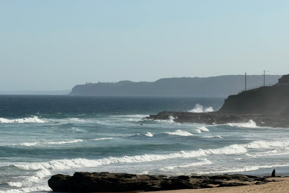 A wide, eye-level shot of a rugged coastline featuring white-capped waves crashing against dark rocky outcrops and a sandy beach under a hazy blue sky.