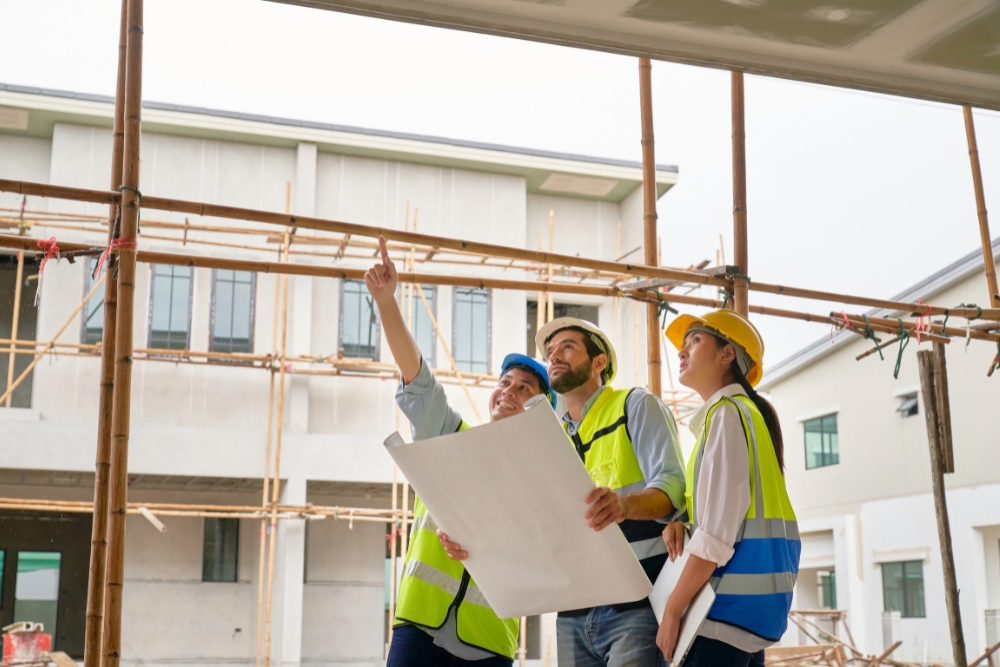 Three construction professionals wearing safety vests and hard hats look upward at a building site.