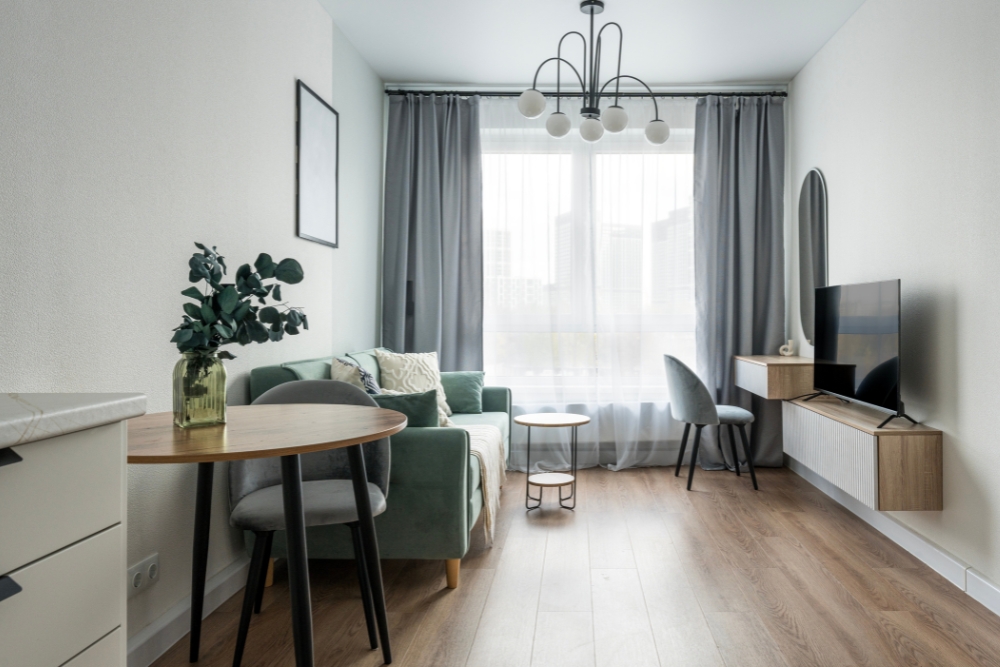 A bright and narrow modern apartment interior featuring a sage green sofa, a small round wooden table, a floating TV console, and a large window with grey drapes.