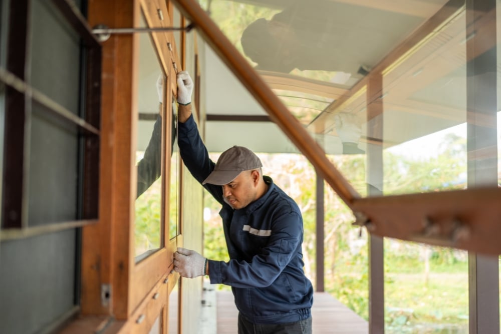 A worker wearing a cap, gloves, and a blue jacket carefully installs or repairs a series of wooden-framed glass windows along an outdoor veranda.