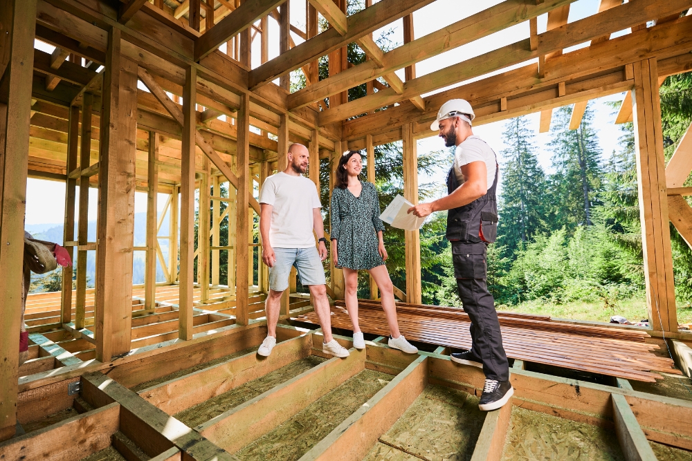 A builder in a hard hat and overalls holds building plans while talking to a couple inside the timber frame of a house under construction.