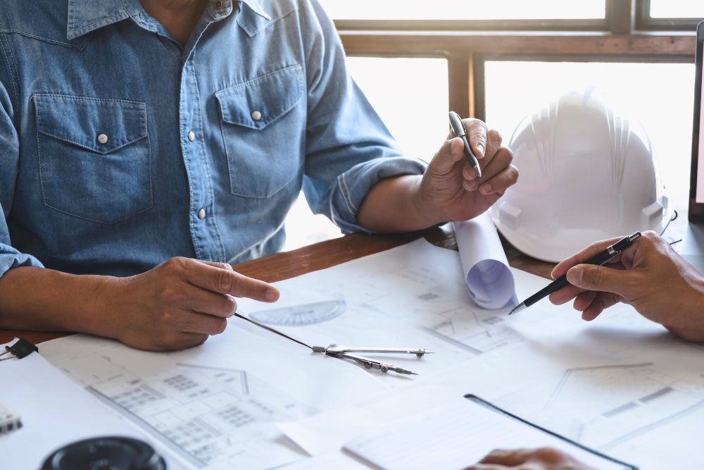 Close-up of two people pointing to architectural blueprints with pens on a wooden table.