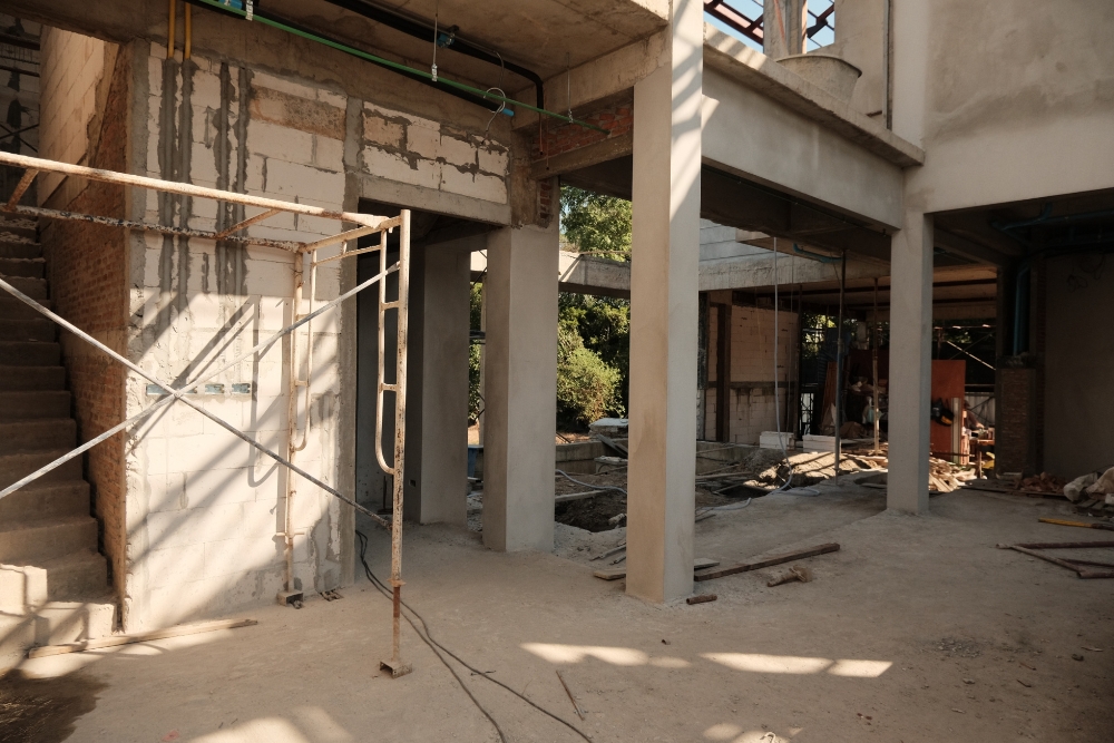 The interior of a multi-storey house under construction, showing bare concrete pillars, unpainted cinder block walls, an unfinished concrete staircase, and metal scaffolding.
