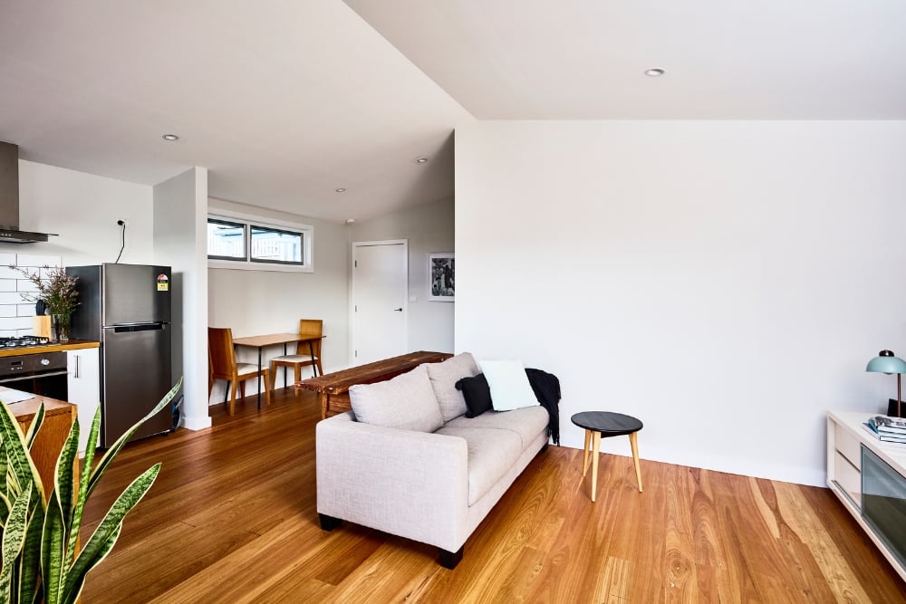 Granny flat interior featuring a light grey couch on polished hardwood floors, opening up to a kitchen with stainless steel appliances and a small dining area.