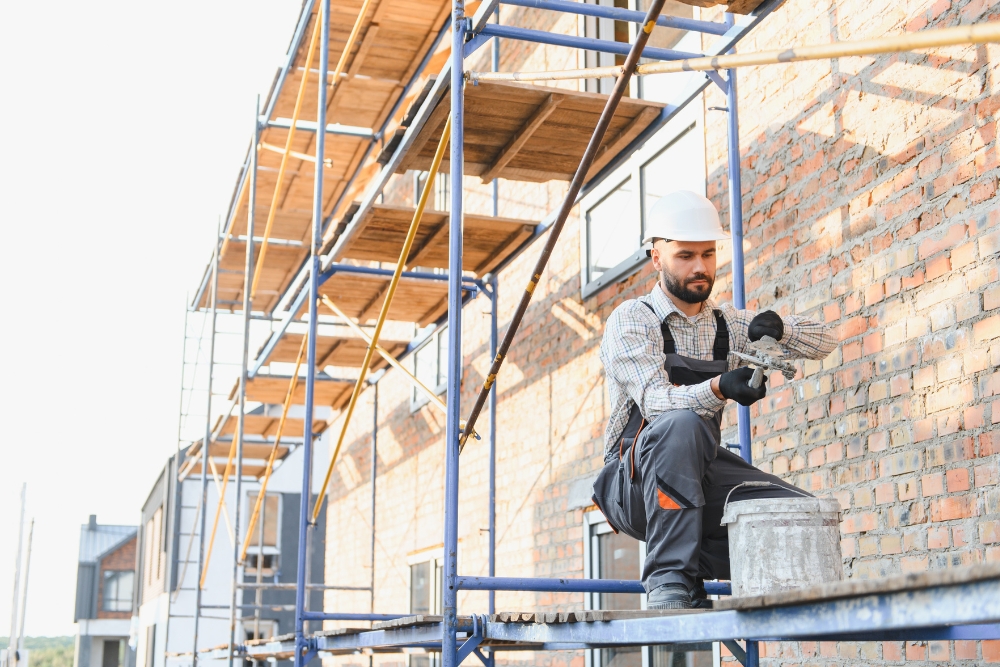 A construction worker wearing a white hard hat and grey overalls kneels on metal scaffolding next to a bucket while preparing mortar on a trowel in front of a brick building.