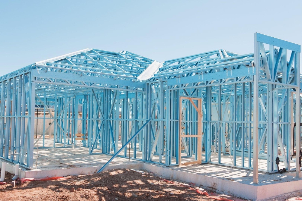 A house under construction showing its light blue steel framing on a concrete foundation against a clear blue sky.