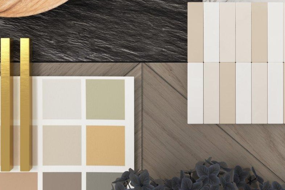 An interior design mood board featuring a paint swatch card, long gold hardware handles, various tile and herringbone wood floor samples, a dark textured stone background, and dark flowers at the bottom.
