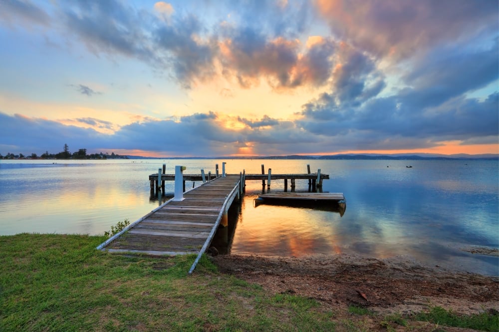 A rustic wooden jetty extending into calm lake waters under a dramatic sunset filled with scattered clouds casting a warm orange and blue glow.