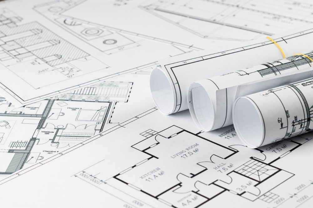 Three rolled-up architectural blueprints resting on a flat, detailed floor plan layout with visible room dimensions and labels.