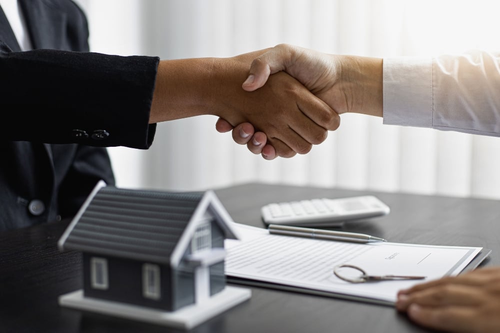 A handshake over a desk featuring a small house model, a contract with a pen, a key, and a calculator.