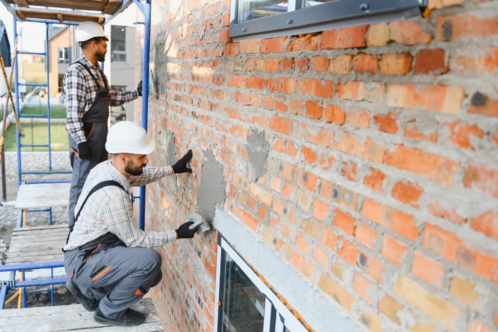 Two construction workers wearing white hard hats and grey overalls stand on metal scaffolding while applying mortar or render to an exterior red brick wall.