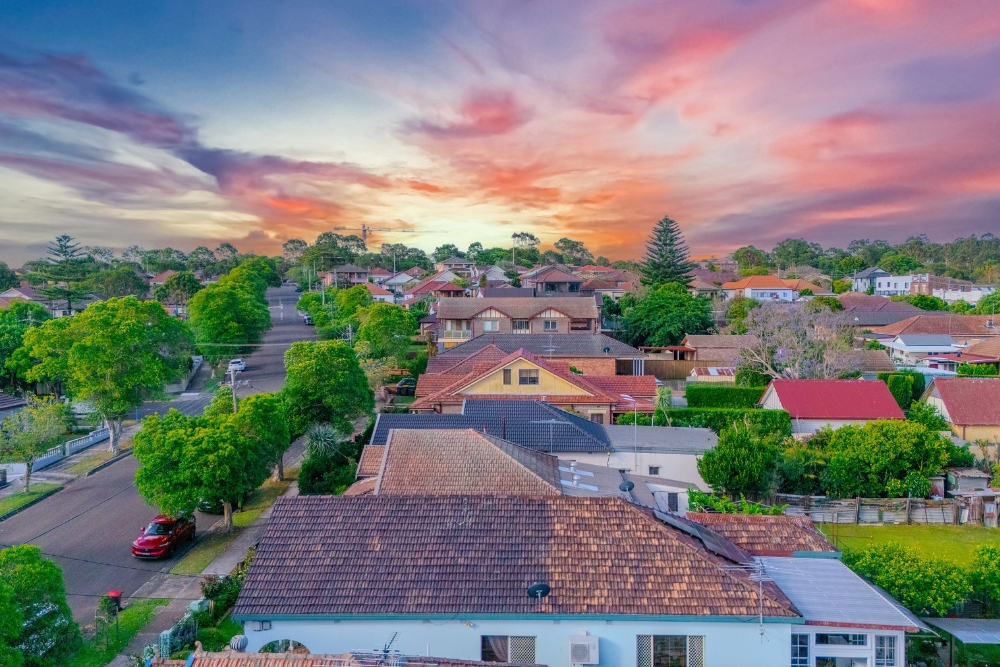 An elevated neighborhood view of suburban homes with tiled roofs and green trees alongside a street, beneath a vibrant sunset with pink, orange, and purple clouds.