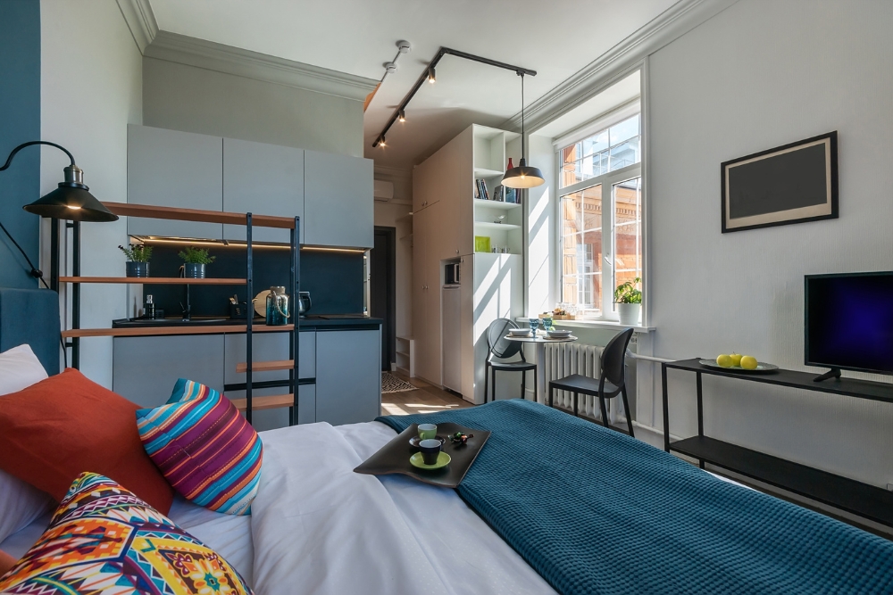 A bed with colourful cushions in the foreground, leading into a compact kitchen area and a small dining table by a window.