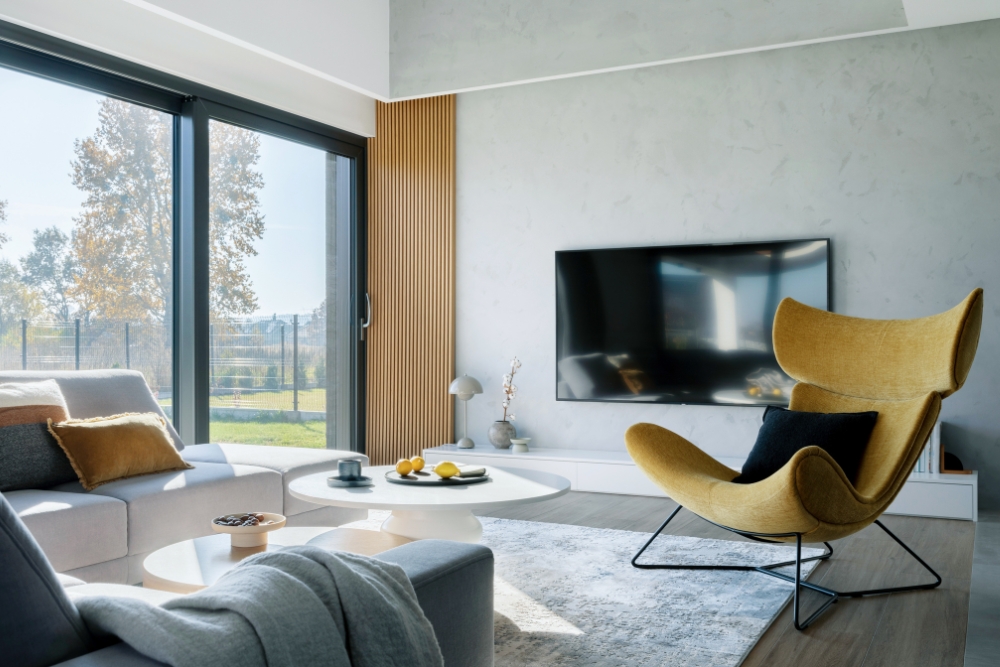 A modern, bright living room featuring a yellow wingback armchair, grey sofas, and large glass doors looking out onto a backyard.