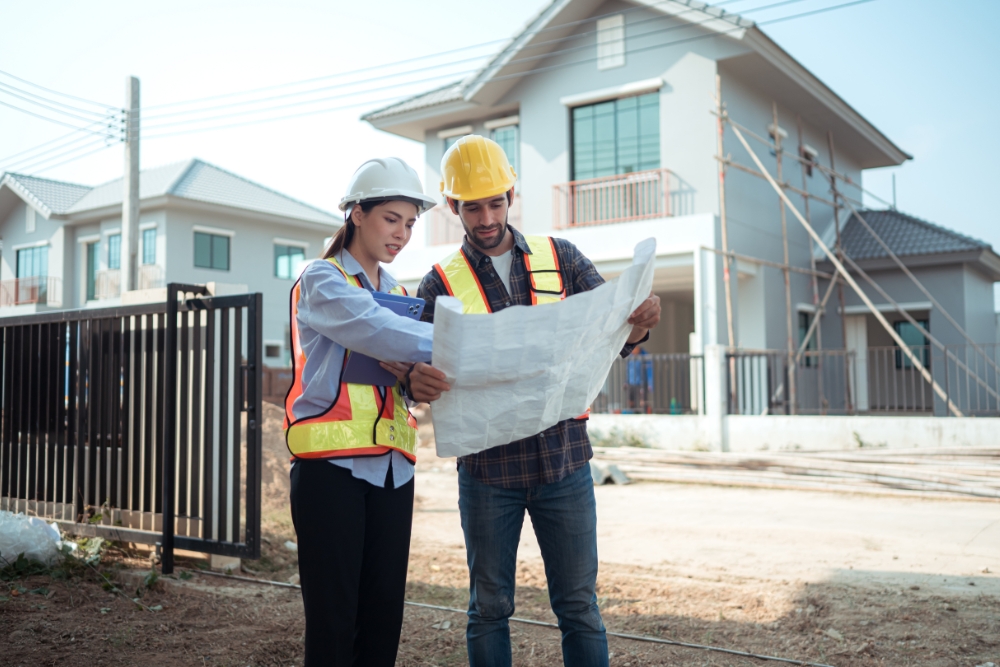 Two construction professionals in safety vests and hard hats review building blueprints outside a modern residential housing development.