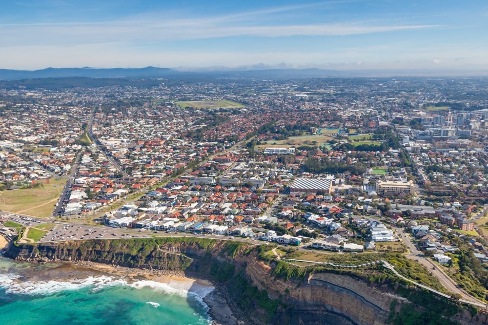 A sprawling coastal city nestled along steep cliffs, meeting a turquoise ocean with white waves crashing on a small sandy beach.
