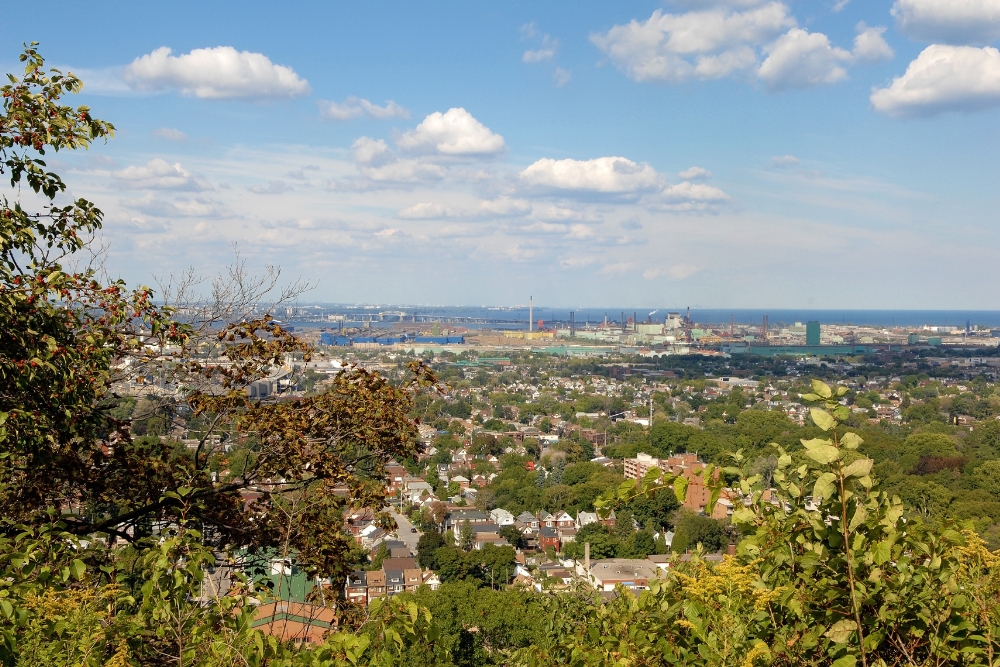 An elevated, panoramic view of the Hamilton East cityscape showing residential neighborhoods, green spaces, and the industrial harbour under a blue, cloudy sky.