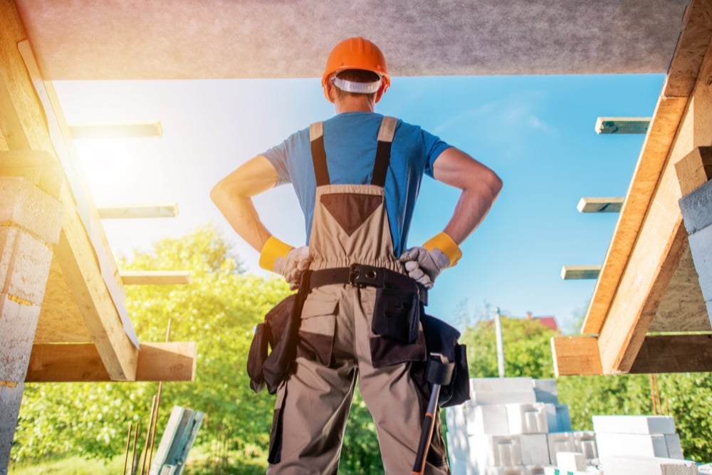 A construction worker wearing an orange hard hat and overalls, standing with hands on hips inside an unfinished structure looking out at a sunny building site.