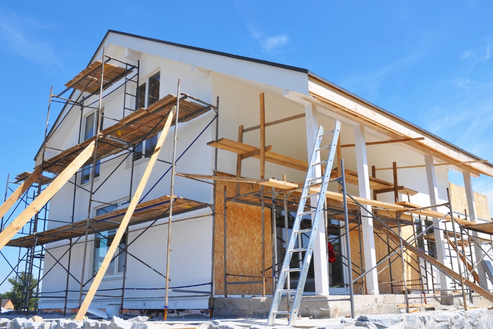 A two-storey white house under construction, surrounded by scaffolding, wooden planks, and an aluminum ladder against a bright blue sky.