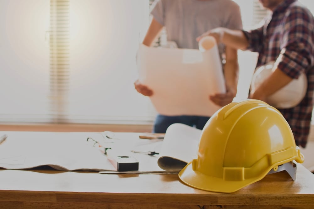A yellow hard hat resting on a wooden desk next to rolled-up blueprints, with two blurry figures holding architectural plans in the background.