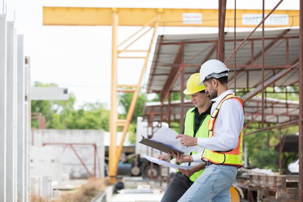 Two construction engineers wearing hard hats reviewing blueprints at an industrial build site.