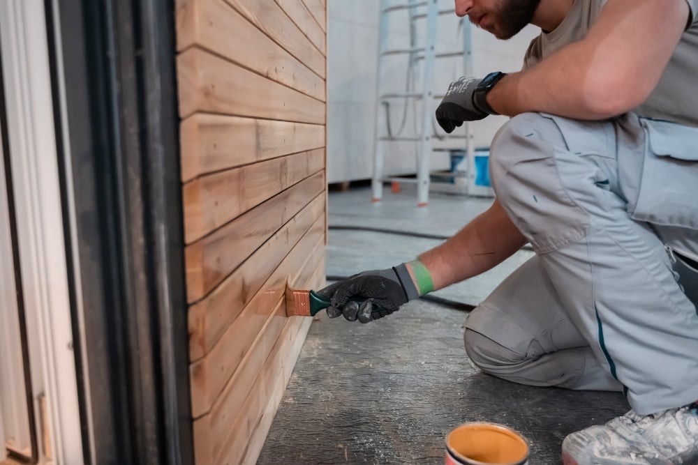 A person applying a clear finish or varnish to a wooden slat wall using a paintbrush.