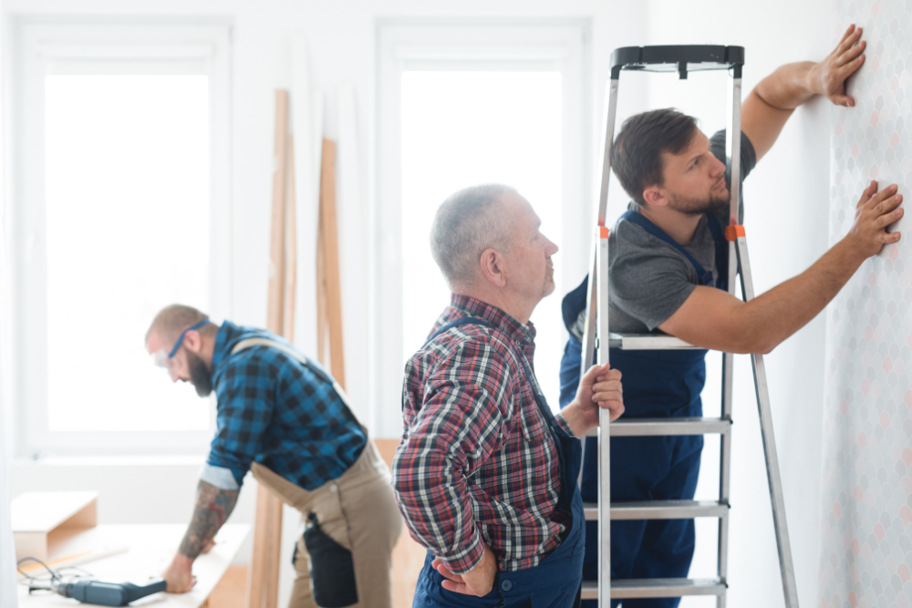 Workers applying patterned wallpaper to a wall during indoor home renovation.