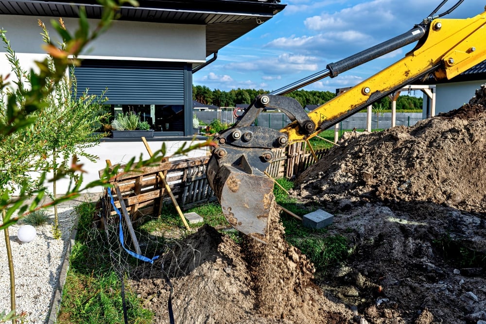 A yellow excavator bucket moving dirt during residential landscaping work outside a house.