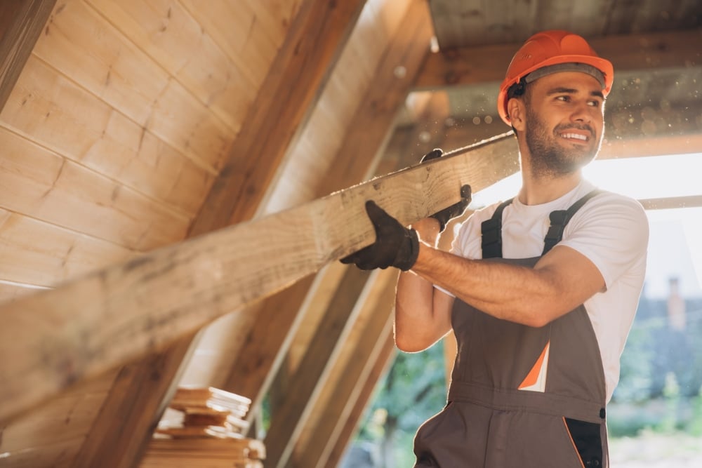 Construction worker in a hard hat carrying a wooden beam inside a timber-frame house.