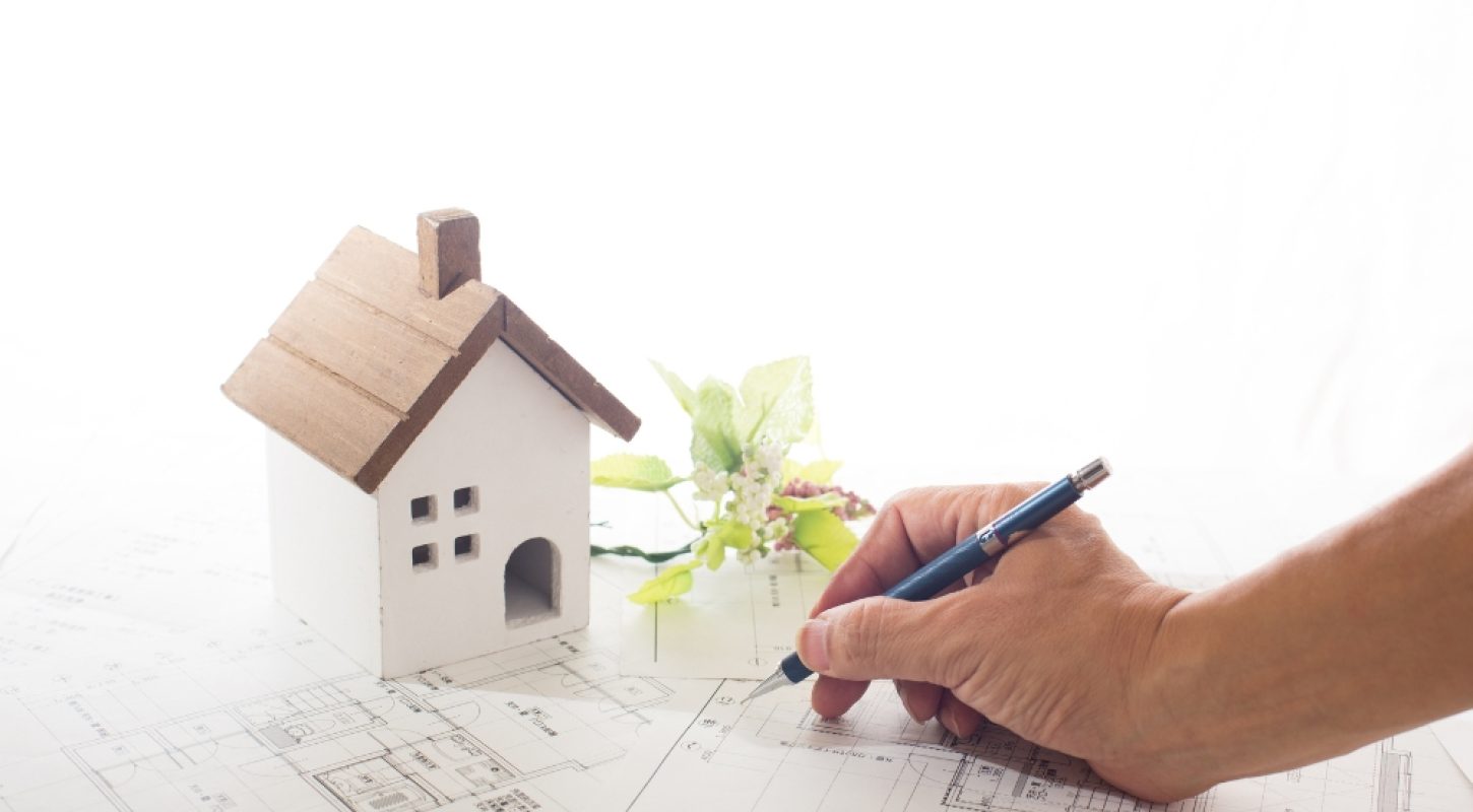 A high-angle, close-up shot of a hand using a mechanical pencil to draft notes on architectural blueprints next to a small, decorative model house.