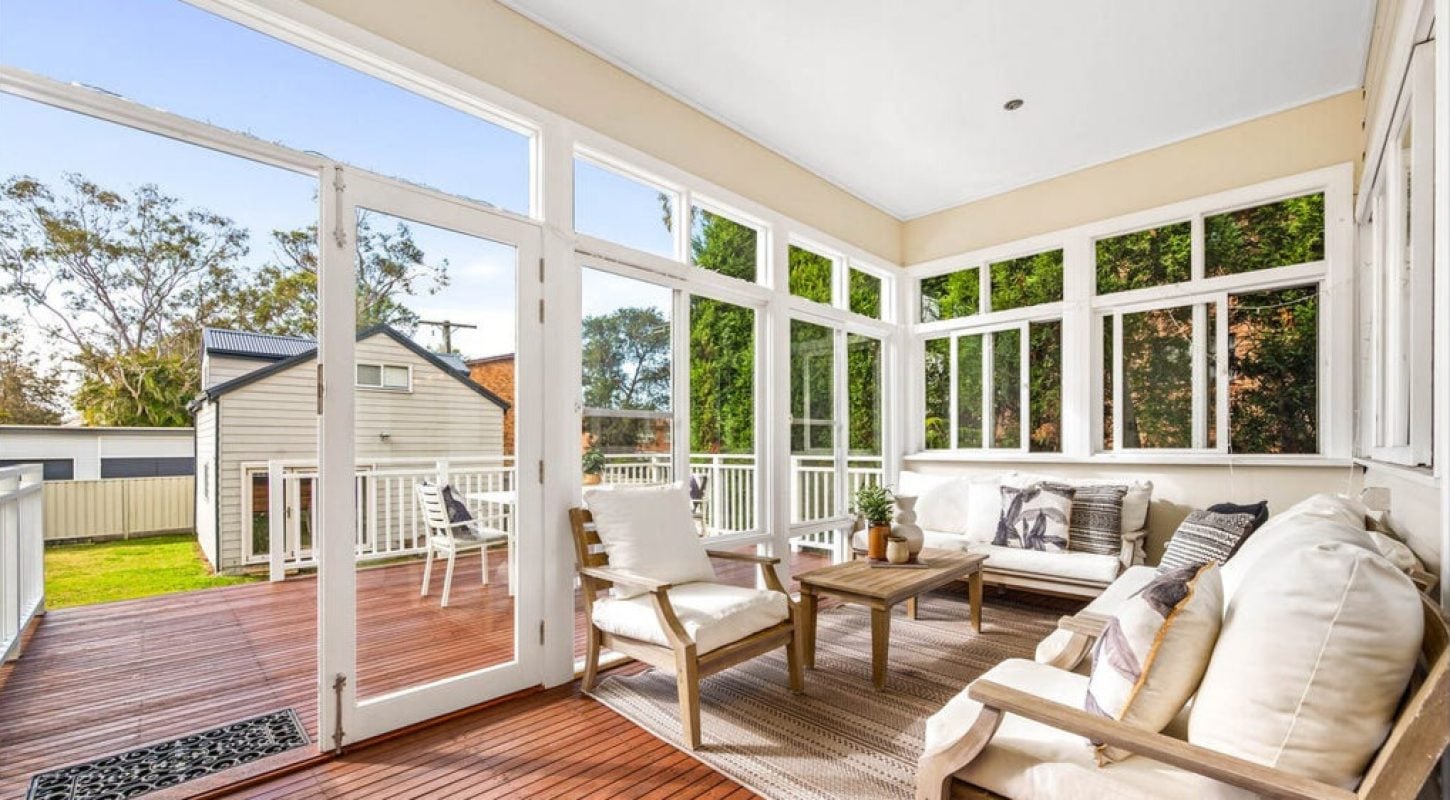 A bright, sun-drenched sunroom featuring white-framed windows and doors that open onto a wooden deck, furnished with comfortable white seating and a neutral area rug.