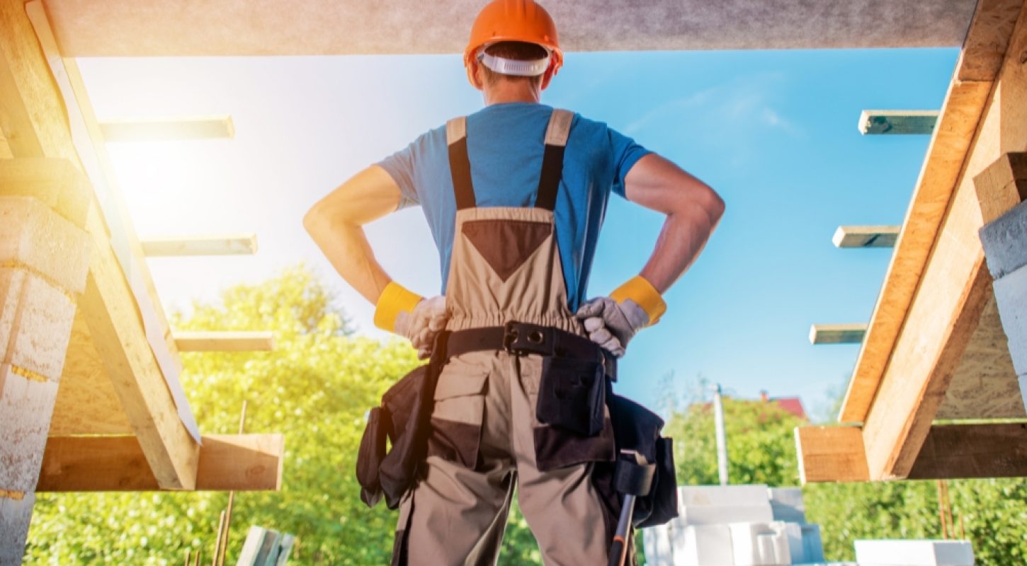 A construction worker wearing an orange hard hat and overalls, standing with hands on hips inside an unfinished structure looking out at a sunny building site.