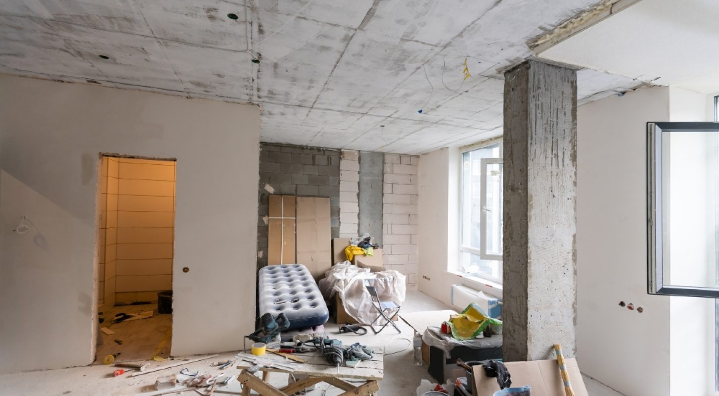 An eye-level shot of a large room in the middle of a renovation, showing exposed concrete ceilings, unfinished block walls, and a variety of construction tools and equipment scattered across the space.